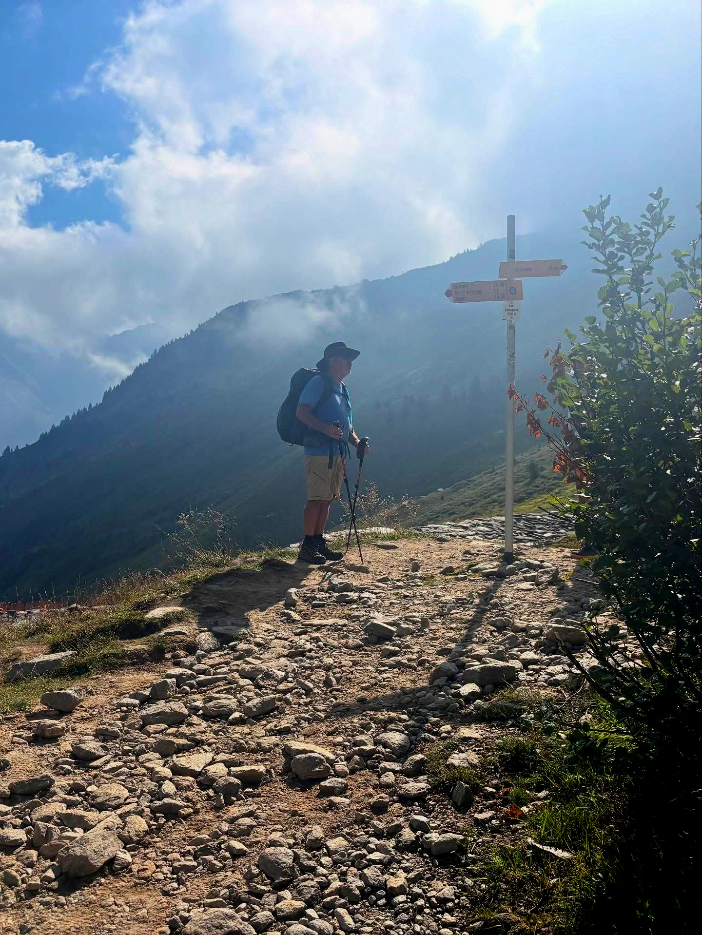 Hiker with a backpack and poles stands on a mountain trail near a signpost, under a partly cloudy sky.