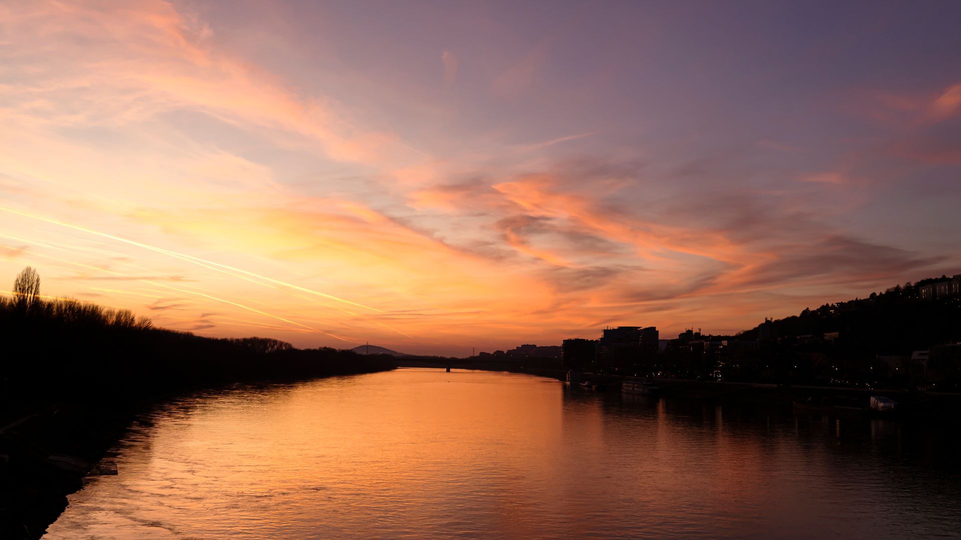 Sunset over a river, with orange and pink sky reflecting in the water. Silhouetted trees and buildings line the banks.