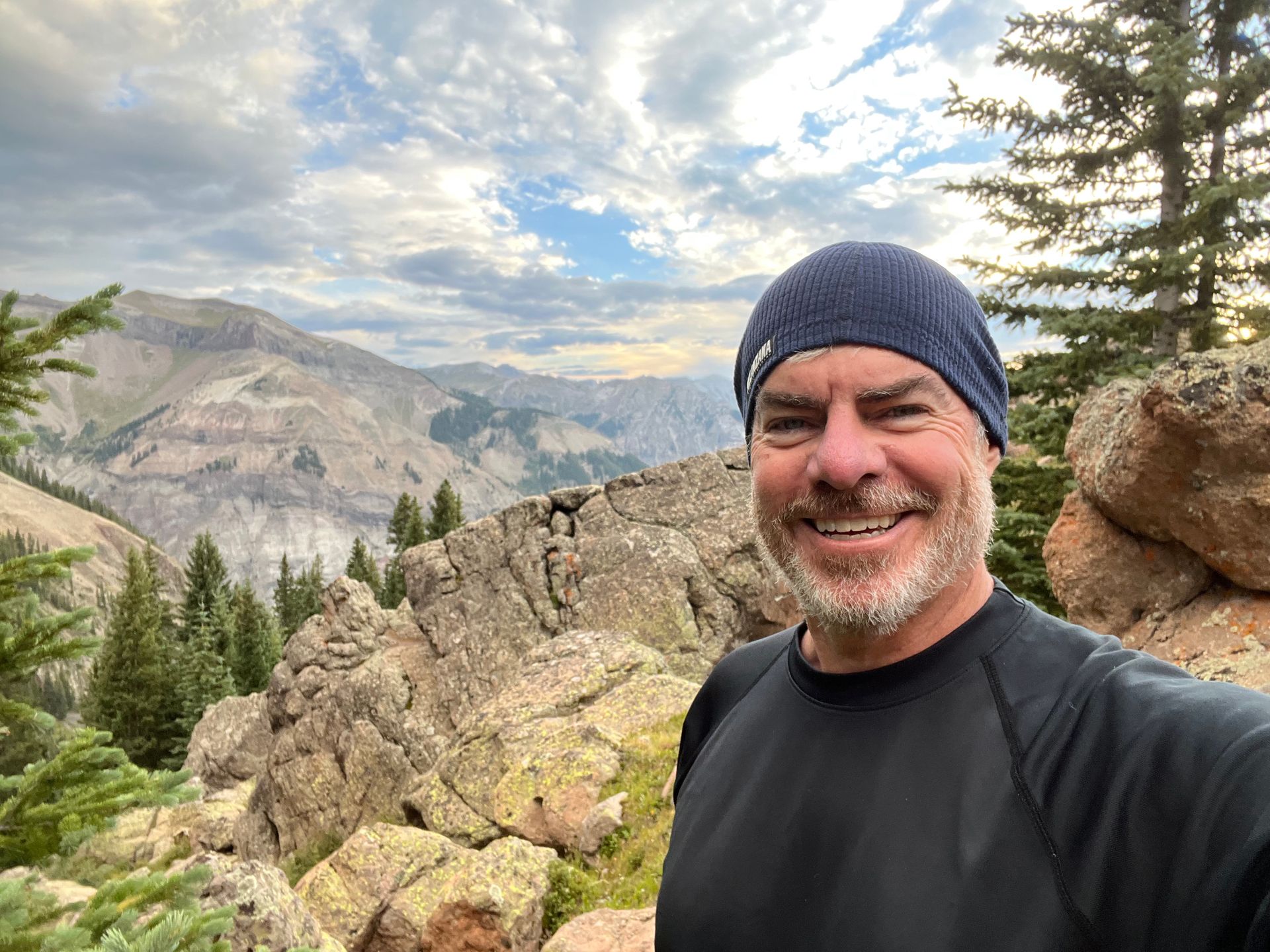 Man smiling, wearing a hat and black shirt, on a mountain with a scenic, cloudy background.