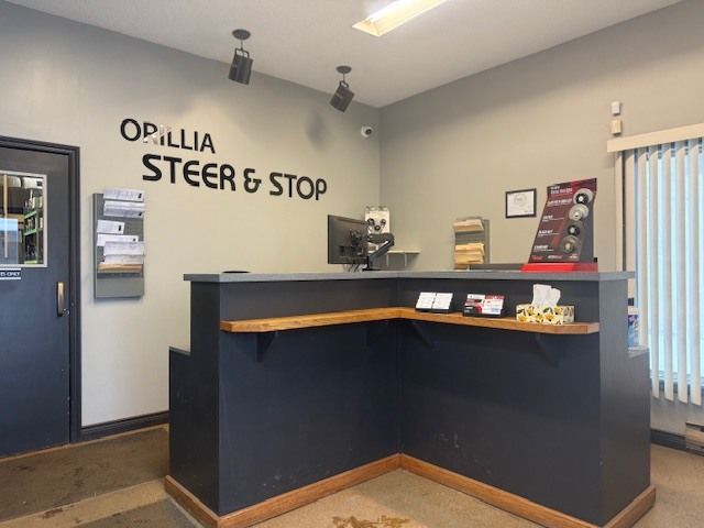 A reception area for Orillia Steer & Stop with an L-shaped dark grey counter, light grey walls, and a door on the left. | Orillia Steer And Stop
