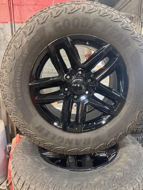 A black 6-spoke truck wheel with a Goodyear Wrangler tire, stacked on top of another tire in a workshop setting. | Orillia Steer And Stop