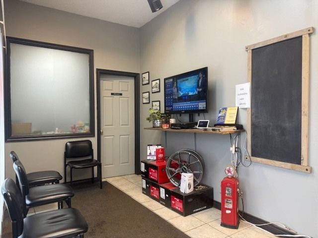 A waiting room featuring three black chairs, a television on a shelf, and a large chalkboard on a light gray wall. | Orillia Steer And Stop