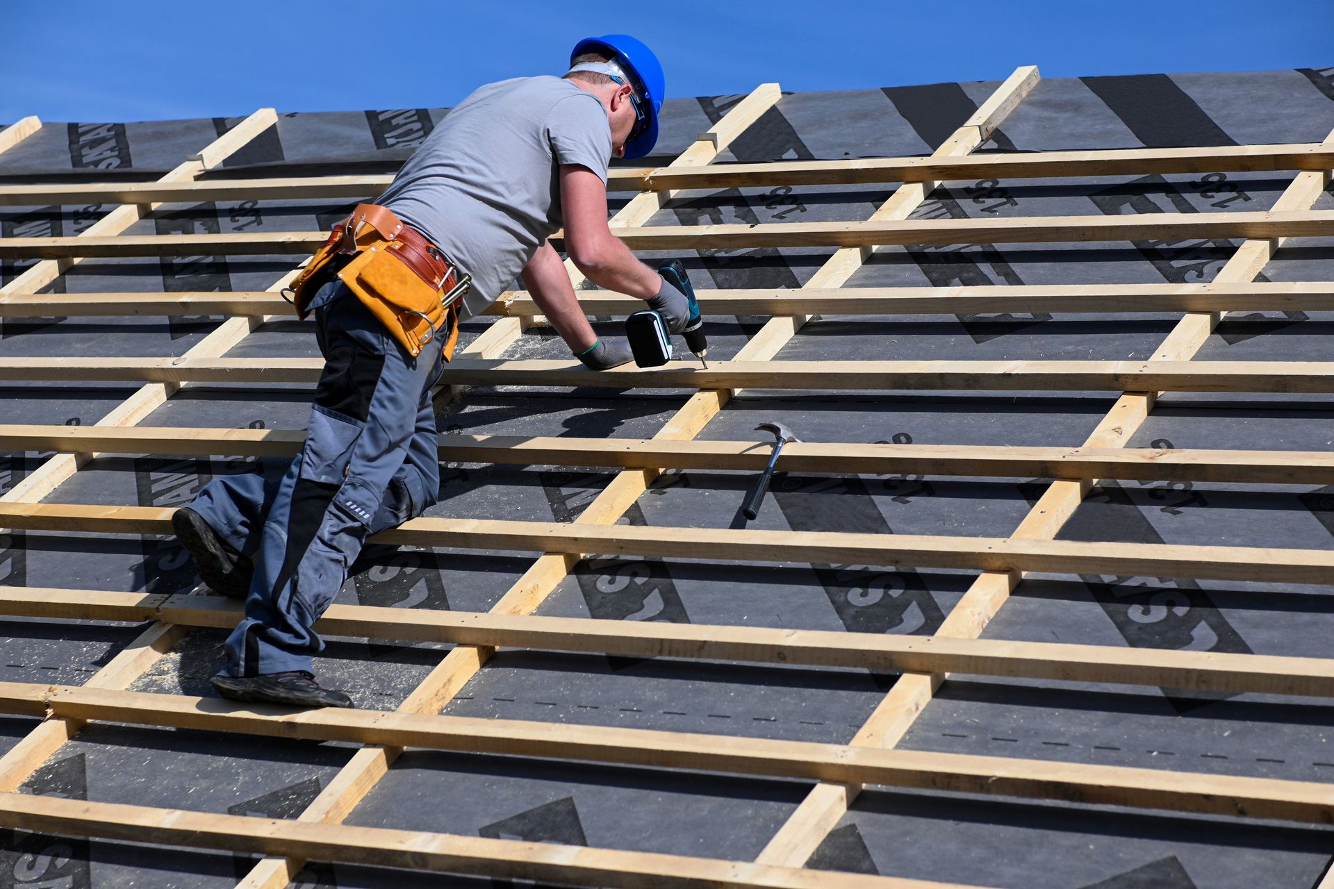 Roofer in hardhat using a tool on a roof under construction; wooden beams and black material visible.