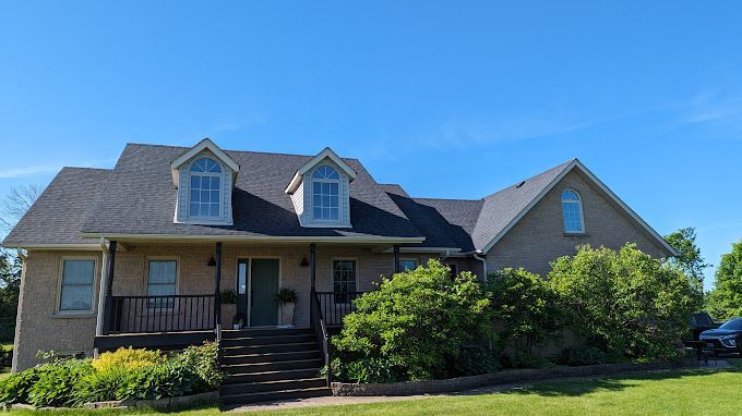 Two-story brick house with dark roof and dormer windows, surrounded by green lawn and trees under a blue sky.
