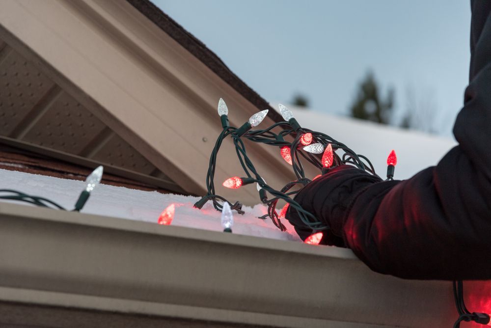 Person in black gloves installing red and white Christmas lights on a roof.