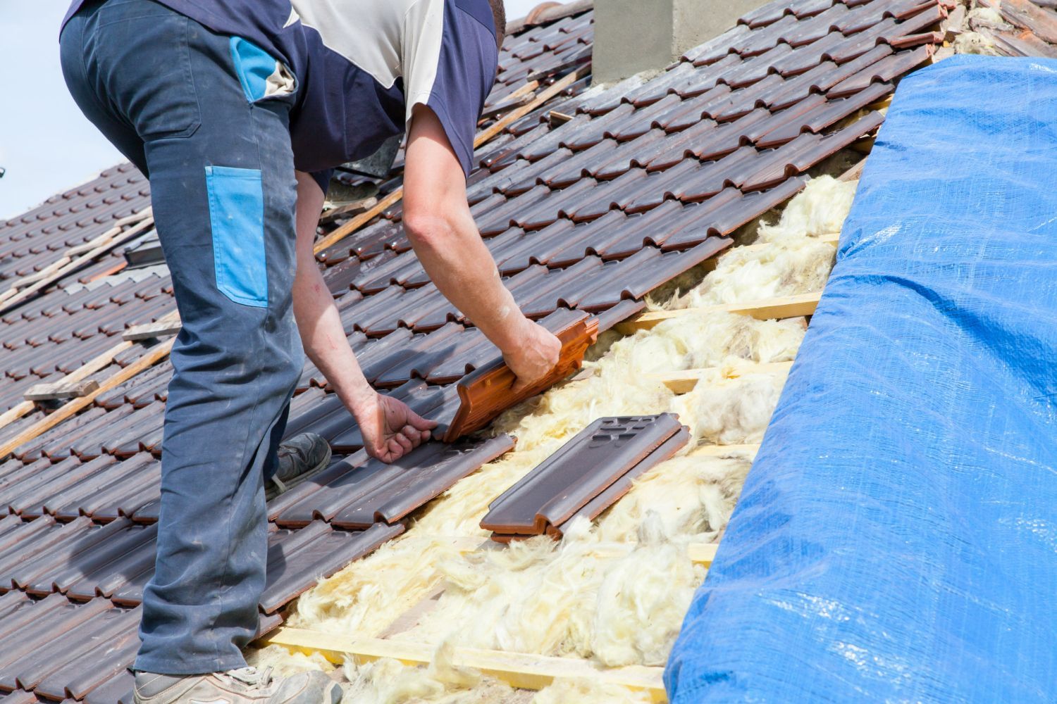 Roofer installs roof tile; blue tarp and insulation visible.