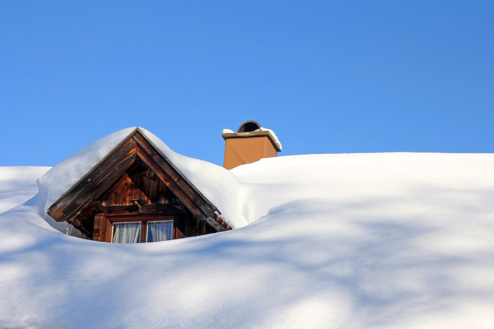 Snow-covered roof with a small wooden dormer and chimney against a bright blue sky.