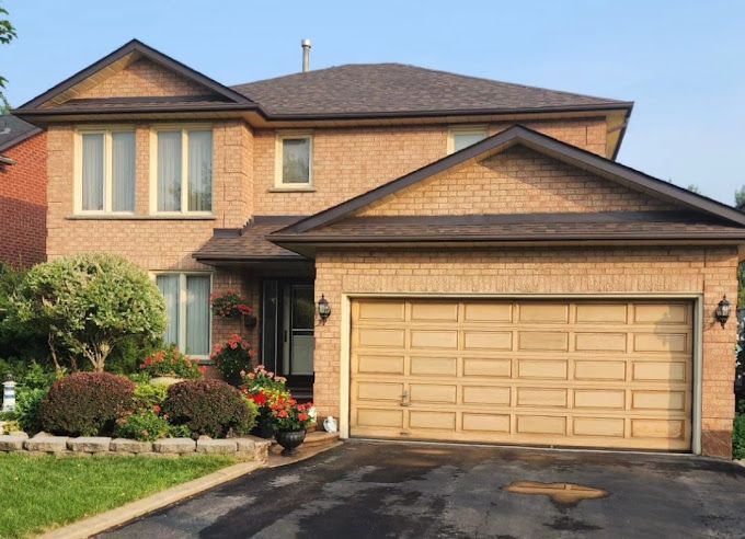Two-story brick house with brown roof and garage door, surrounded by green shrubs and a black driveway.