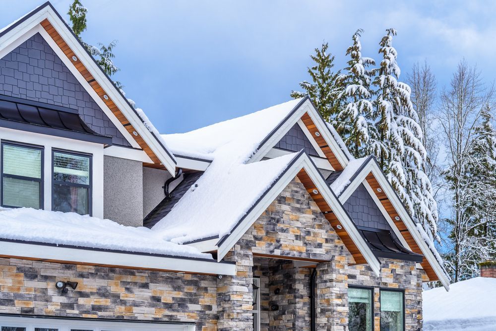 Snow-covered home with stone facade and dark roof trim against a backdrop of snow-covered trees.