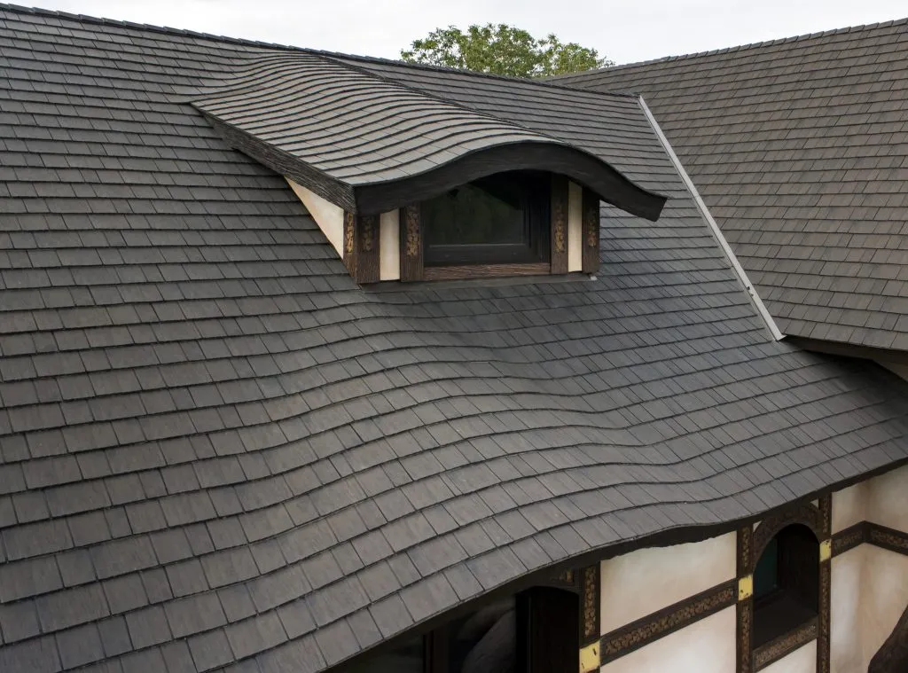 Black shingled roof with a curved dormer window on a building with a white and brown exterior.