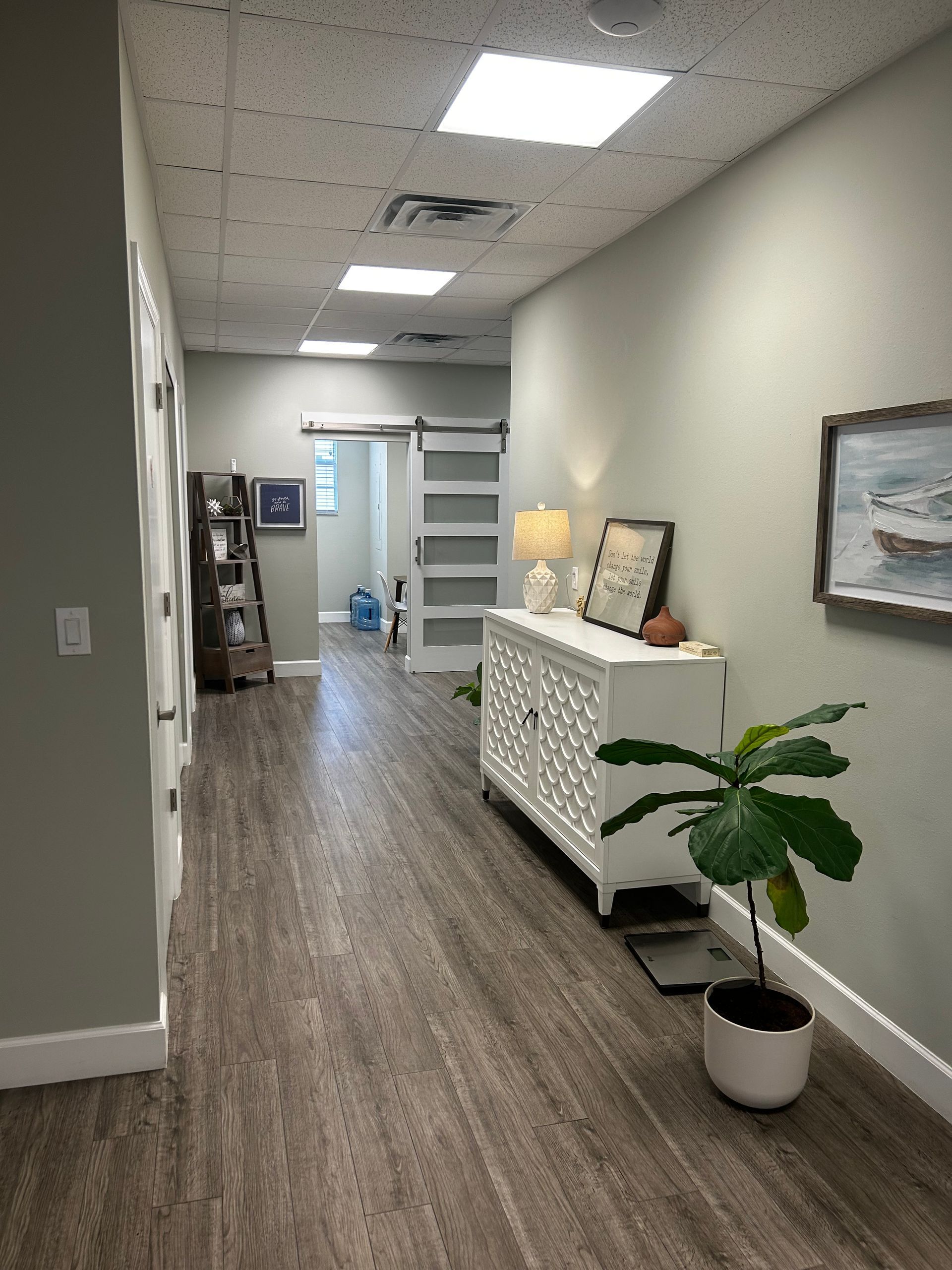 Quiet office hallway with wood floors, white walls, framed art, and a potted plant near a console table.