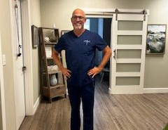 Man in navy scrubs standing with hands on hips in a bright hallway, smiling.