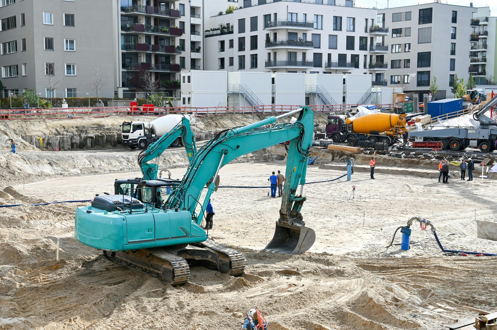 Turquoise excavator at a muddy construction site with apartment buildings and workers in the background