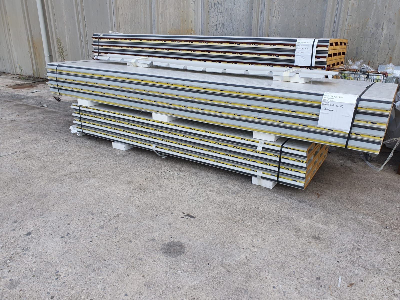 Pile di pannelli isolanti per pareti, grigi e gialli, su pallet all'esterno di un edificio.