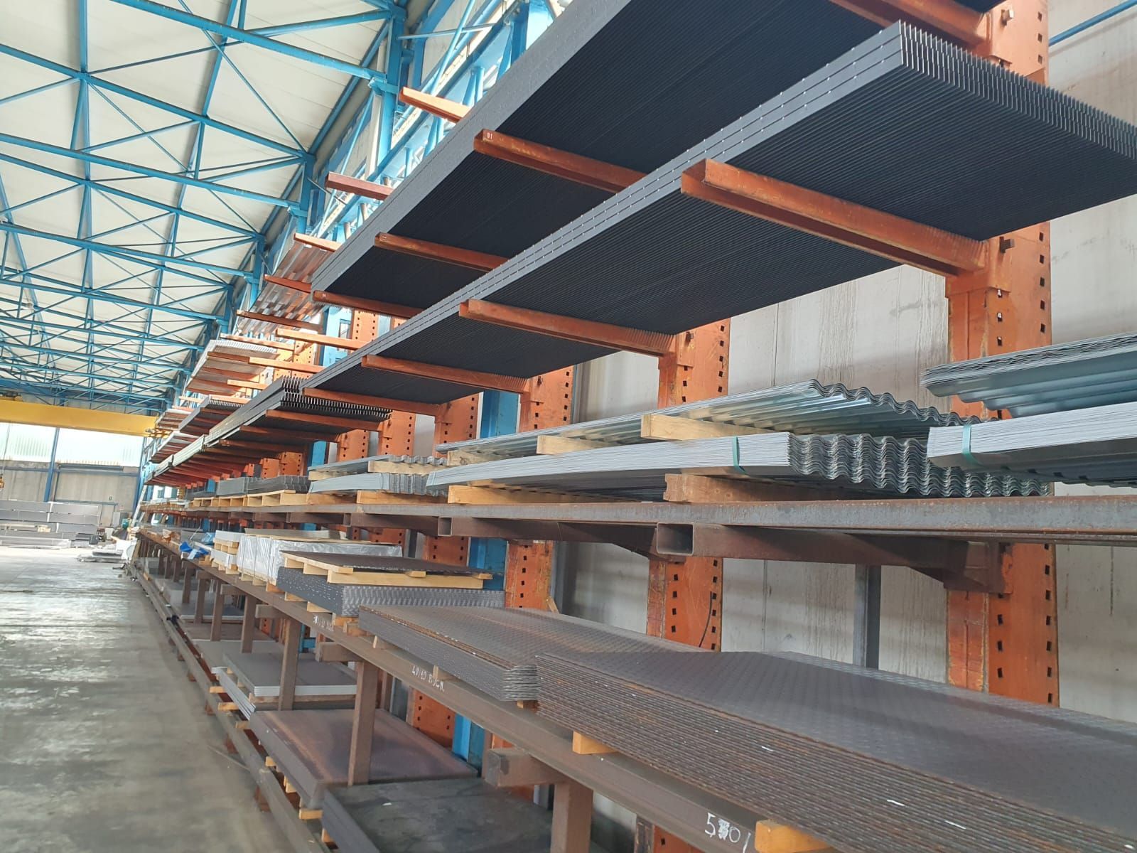 Scaffalatura di magazzino piena di varie lamiere e barre; supporti arancioni, soffitto blu.