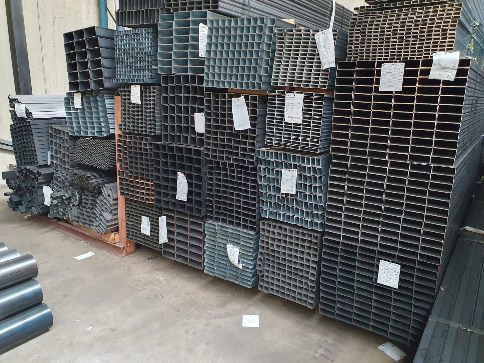 Pile di vari profili in acciaio, tra cui tubi quadrati e rettangolari, in un magazzino.