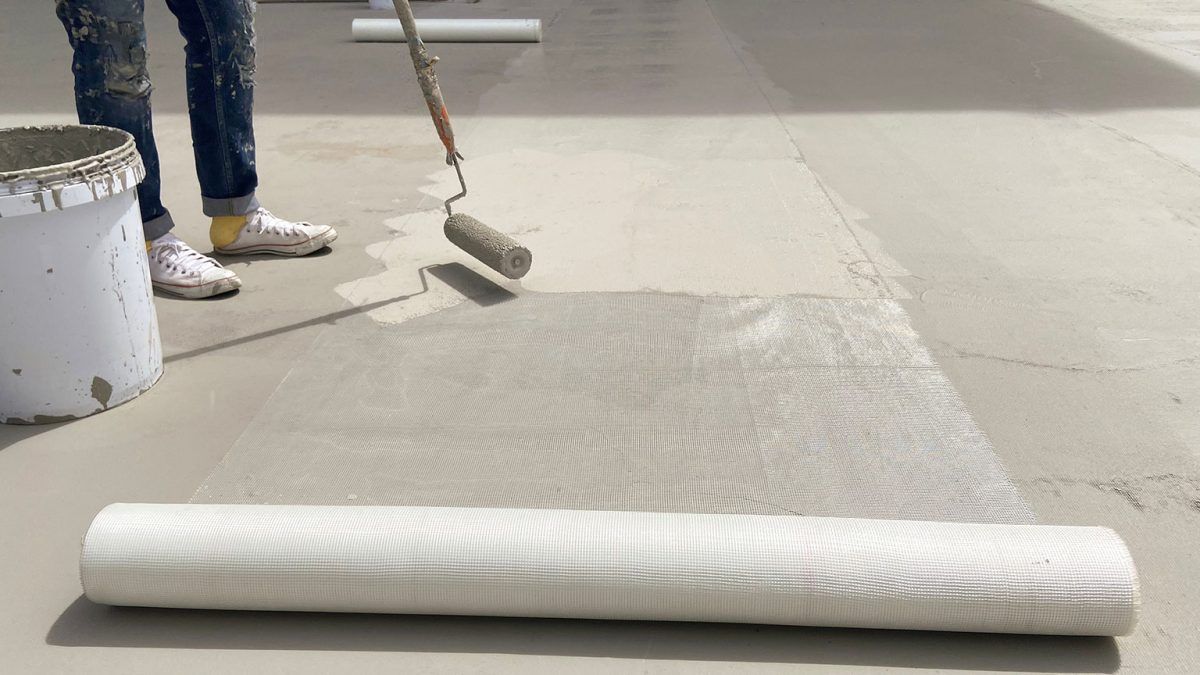 Person using a roller to apply a gray coating to a concrete floor, next to a roll of white fabric.