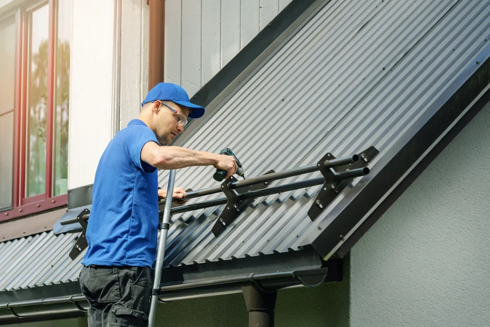 Man in blue shirt and cap installing metal roof brackets on a house.