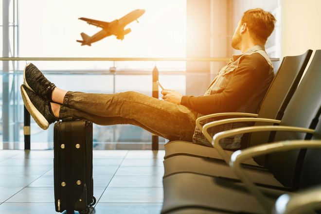 A man is sitting in an airport waiting area with his feet on a suitcase.