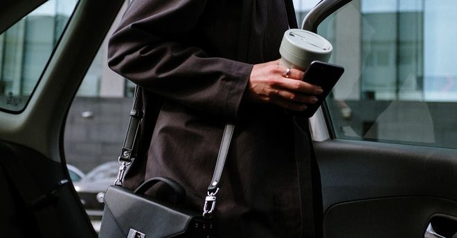 A woman is standing in a car holding a cup of coffee and a cell phone.