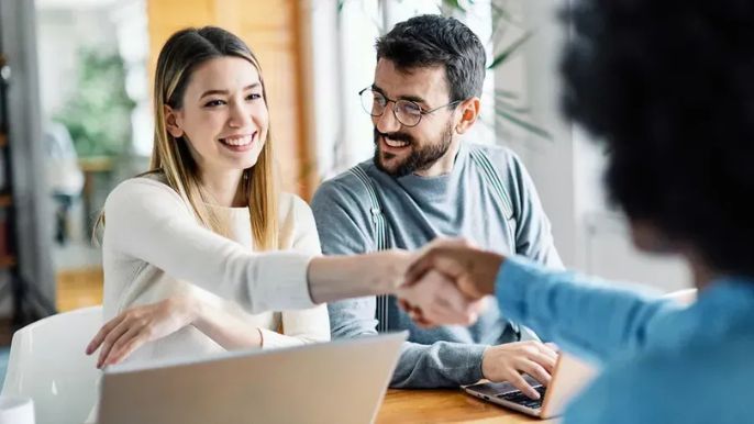 A man and a woman are shaking hands while sitting at a table with a laptop.