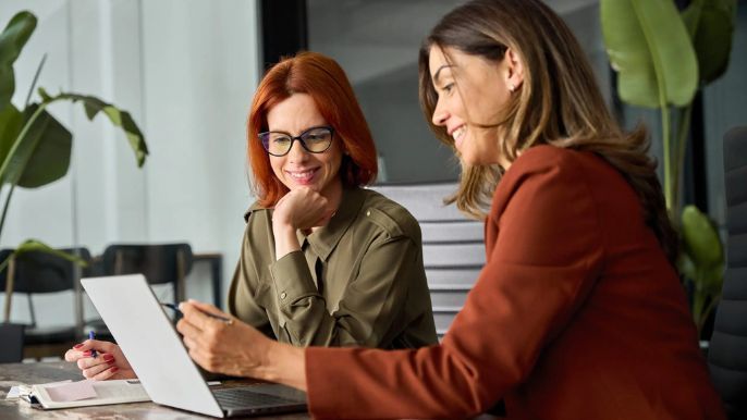 Two women are sitting at a table looking at a laptop.