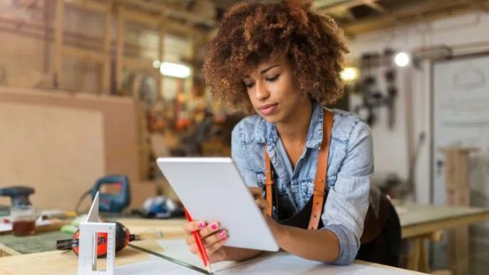 A woman is sitting at a table in a workshop using a tablet computer.