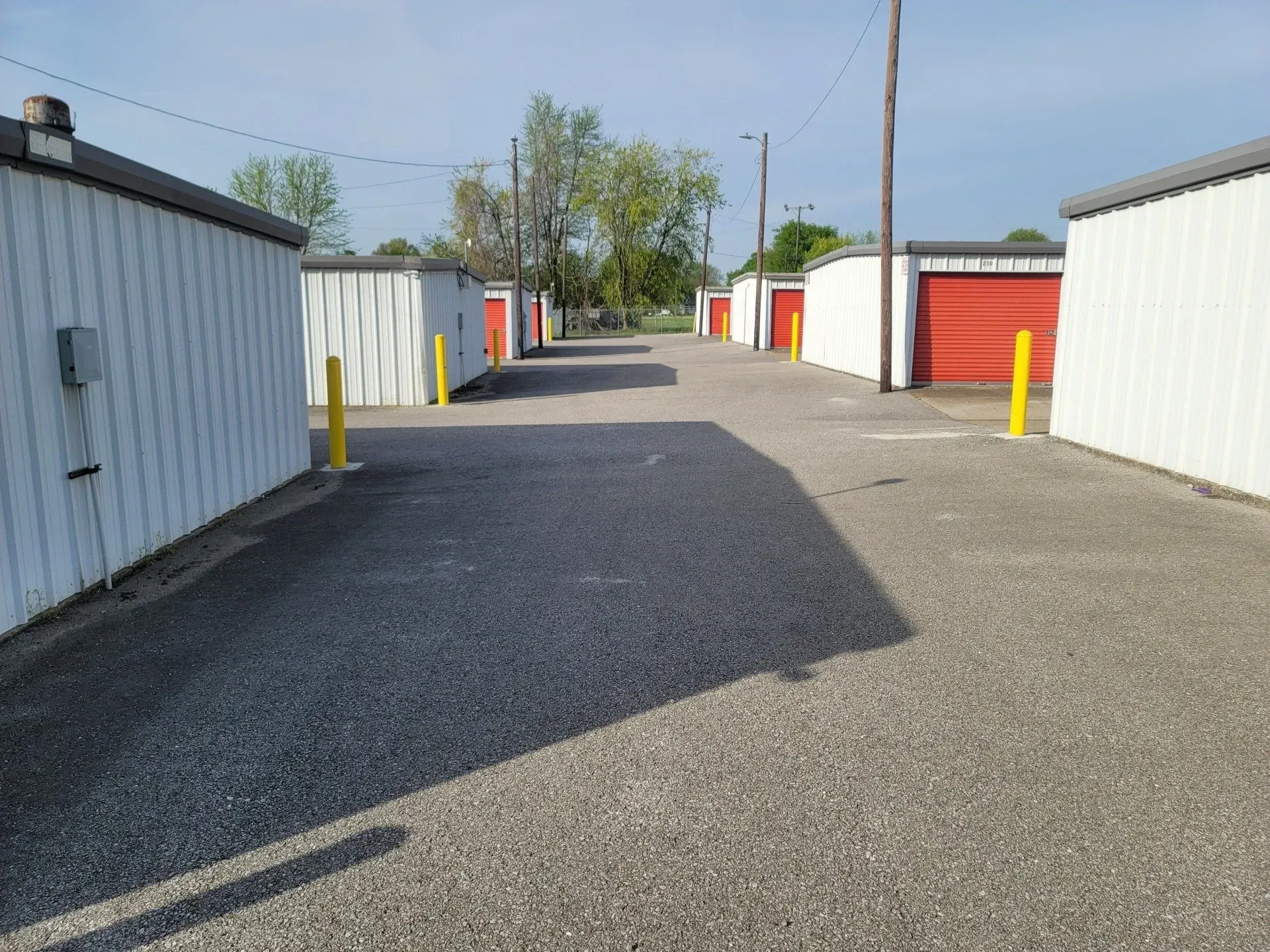 Storage units with red doors line a gravel driveway, yellow posts, sunny day.