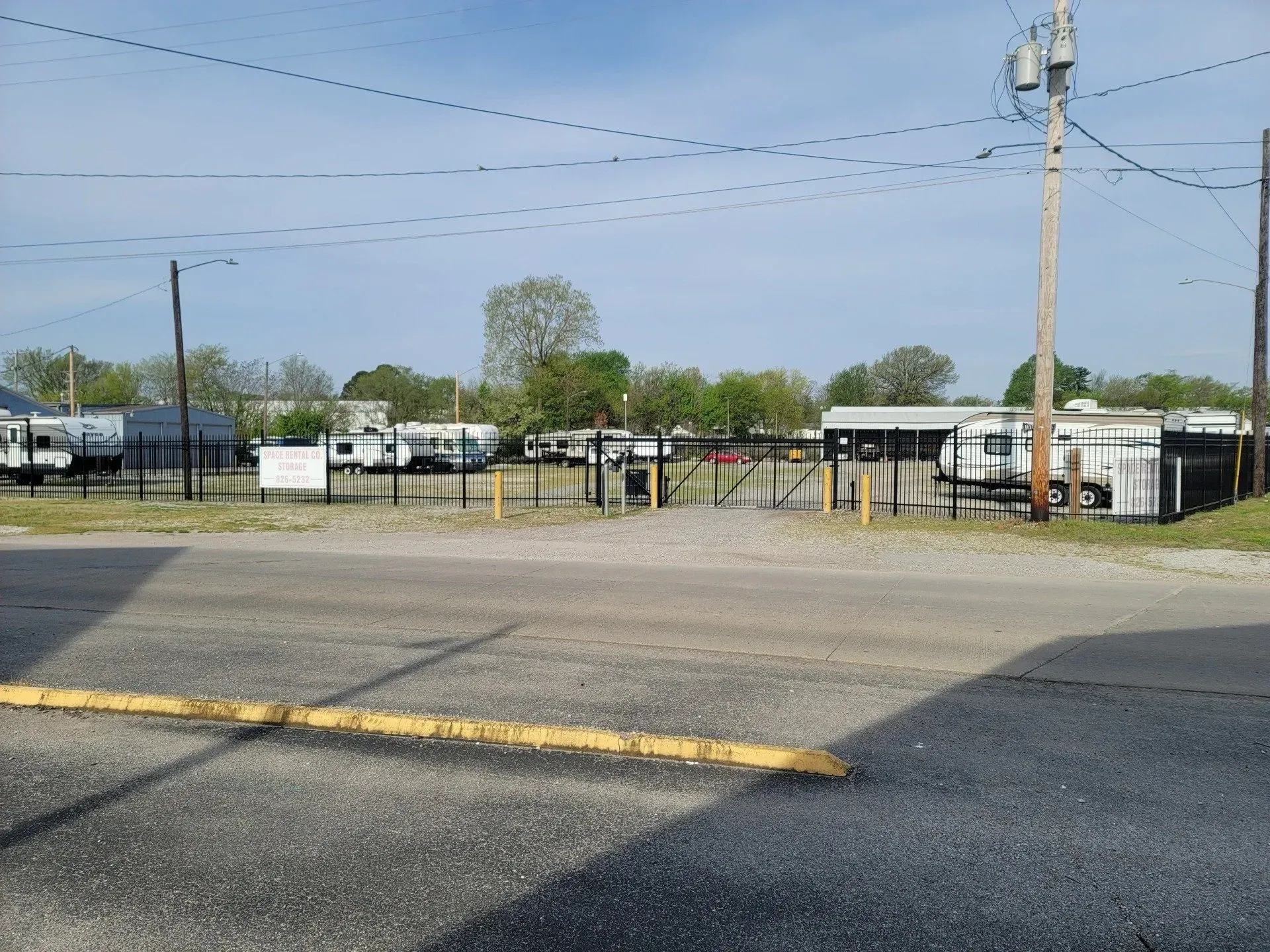 Gated storage facility entrance with parked trailers and parked vehicles; asphalt in the foreground.