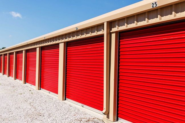 Red storage unit doors in a row, beige trim, gravel ground, bright blue sky.