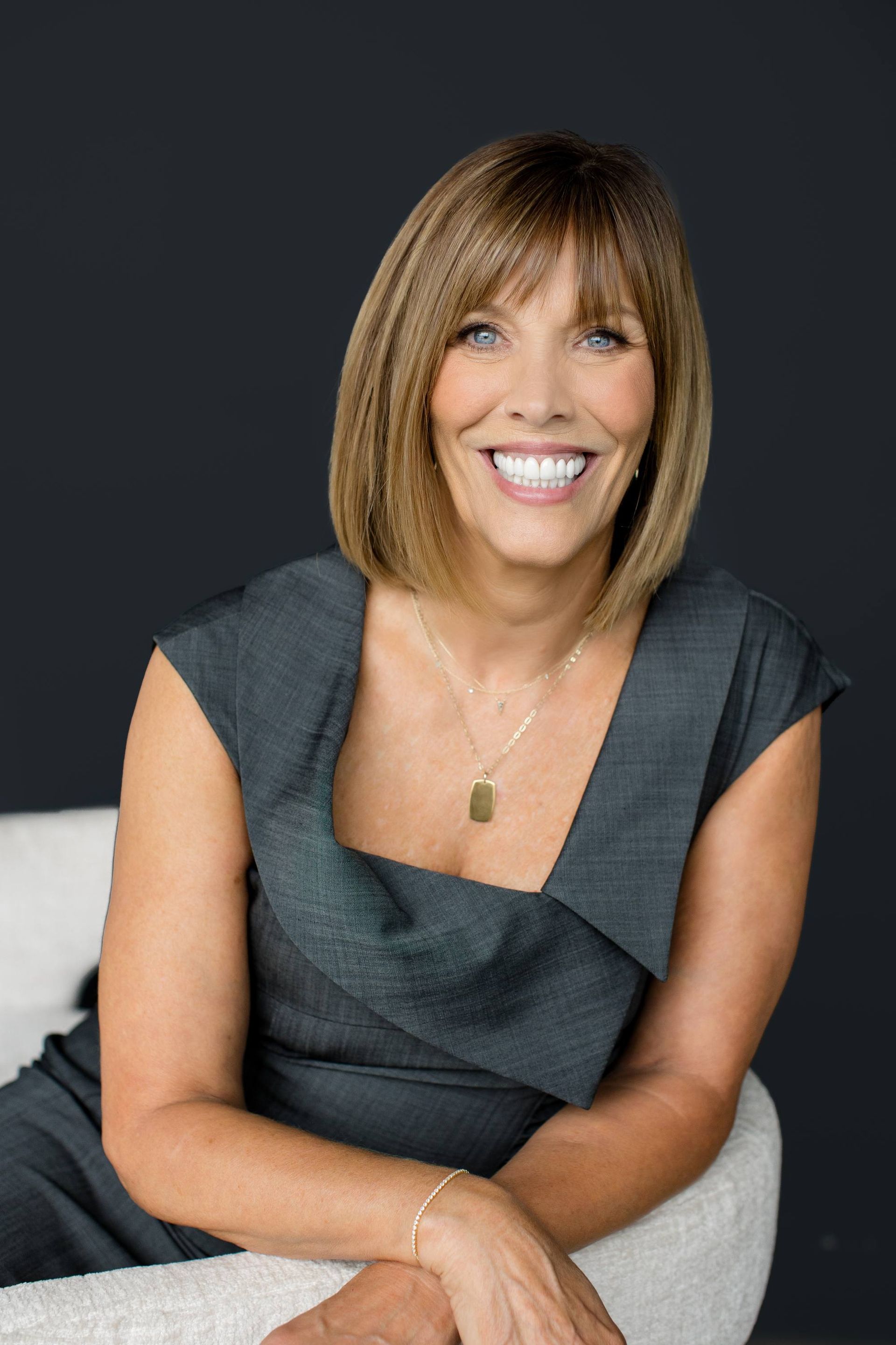Woman with short brown hair, smiling, wearing a gray dress, sitting in a chair.