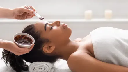 Person receiving a facial mask in a spa; a hand applies mask to forehead.