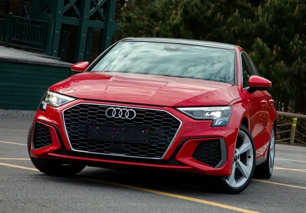 Red Audi sedan parked on a city street, front view, glossy red paint and black grille.