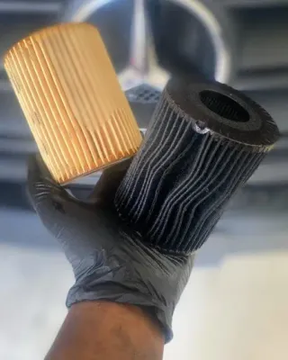 Gloved hand holding two pleated filters, one tan and one black, in a car engine bay