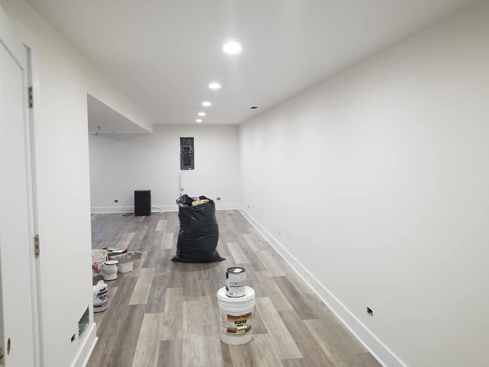 Empty long white room with gray wood flooring, paint supplies, and recessed ceiling lights