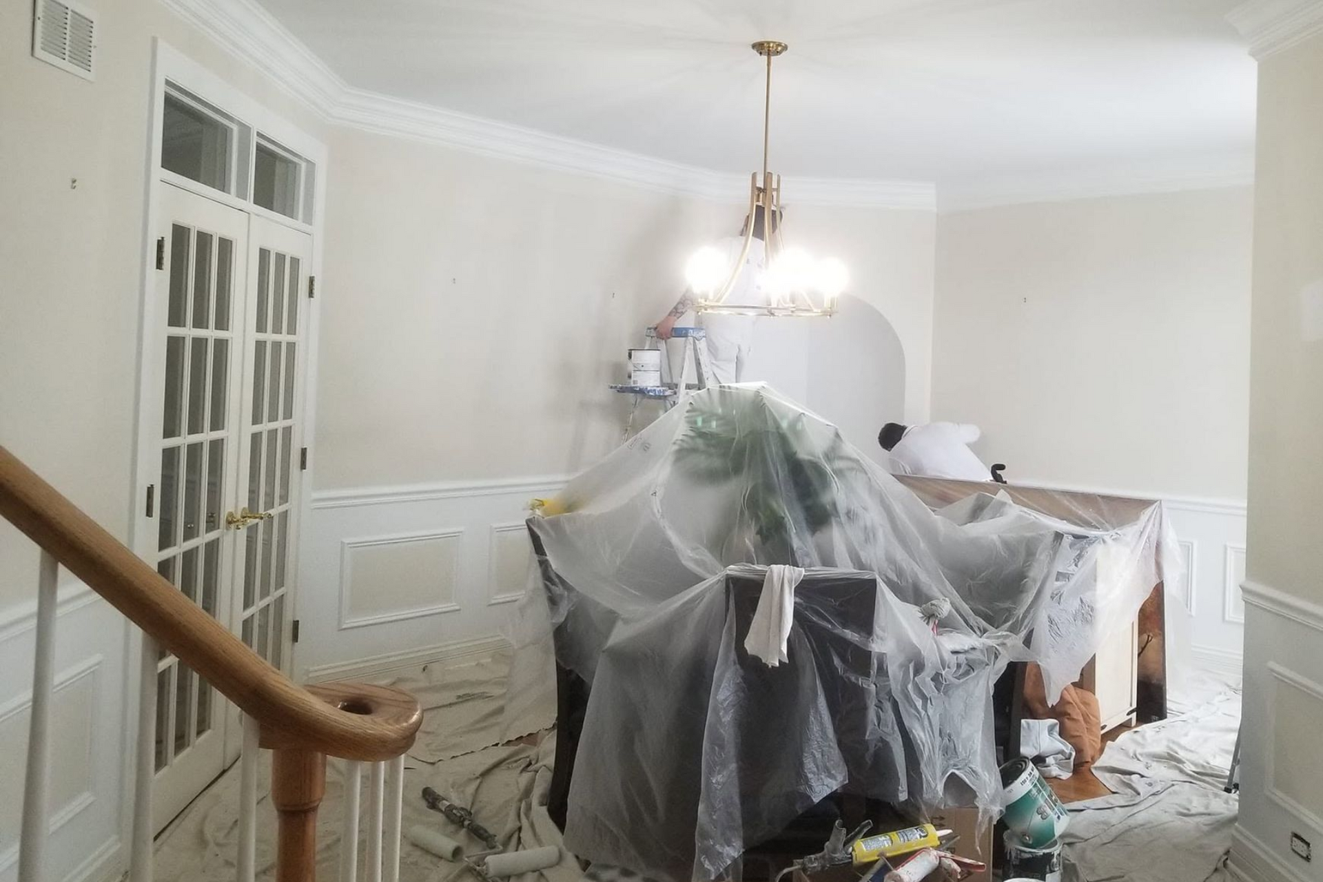 Dining room covered in plastic during renovation, with chandelier, white walls, and a staircase railing in view