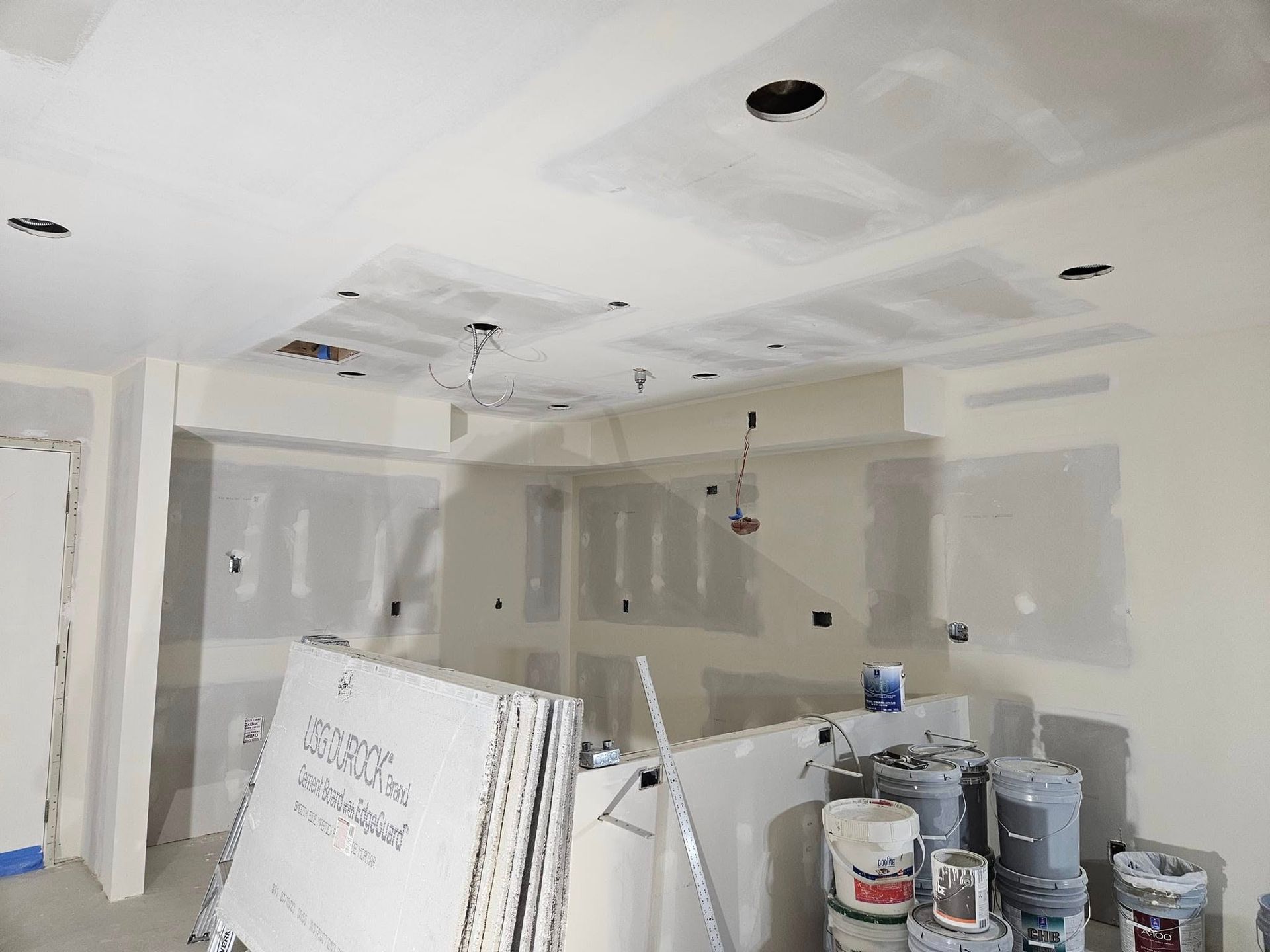 Unfinished room with drywall patches, ceiling lights, and stacks of paint and construction materials