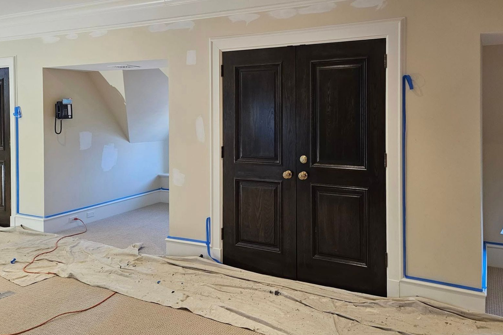 Interior hallway with dark double doors, beige walls, and blue painter’s tape along the trim and floor.