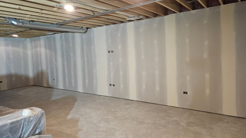 Unfinished basement with exposed ceiling joists and white wall panels, viewed across a concrete floor.