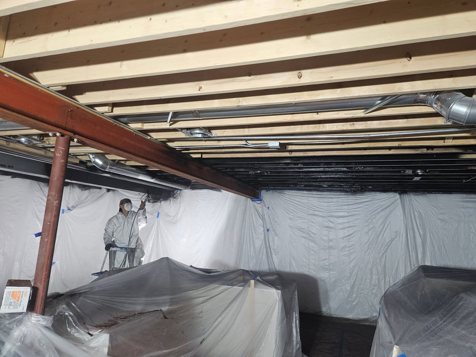 Basement with exposed beams, plastic-covered furniture, and a person standing near the back wall.