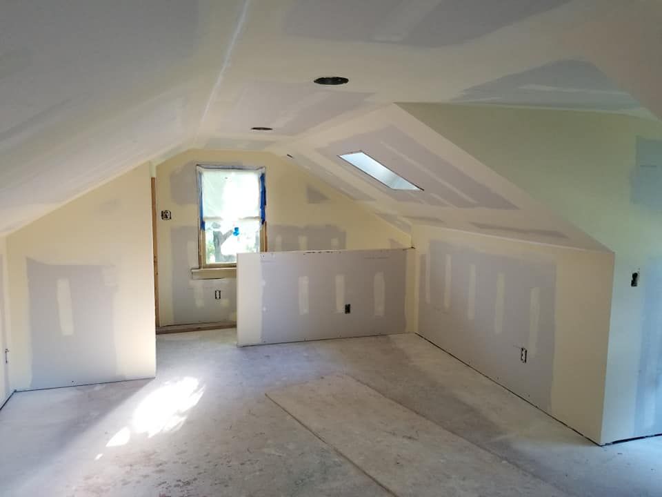 Unfinished attic room with white drywall, sloped ceiling, and a small window letting in sunlight.