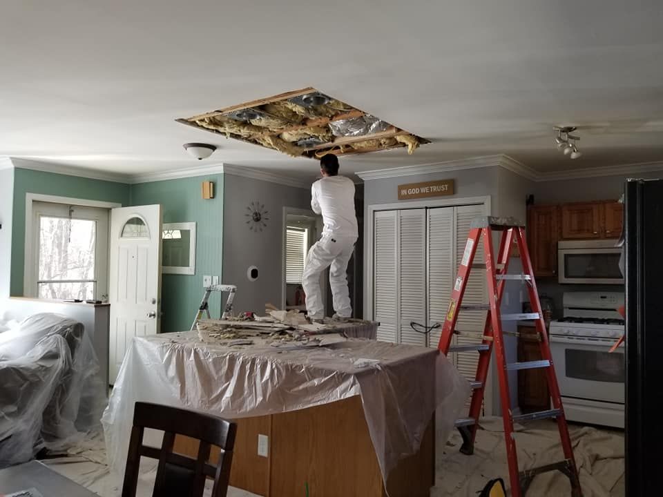 Worker on a ladder repairing a damaged kitchen ceiling, with tools, plastic-covered furniture, and a red step ladder.