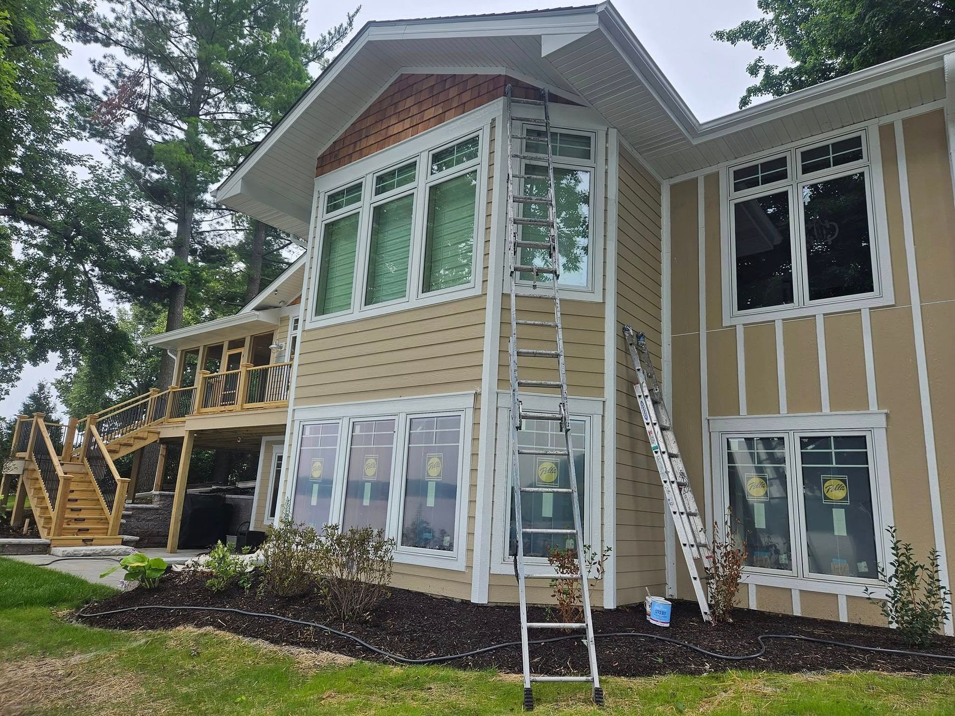 Two-story beige house with white trim, large windows, and wooden stairs under construction in a wooded yard
