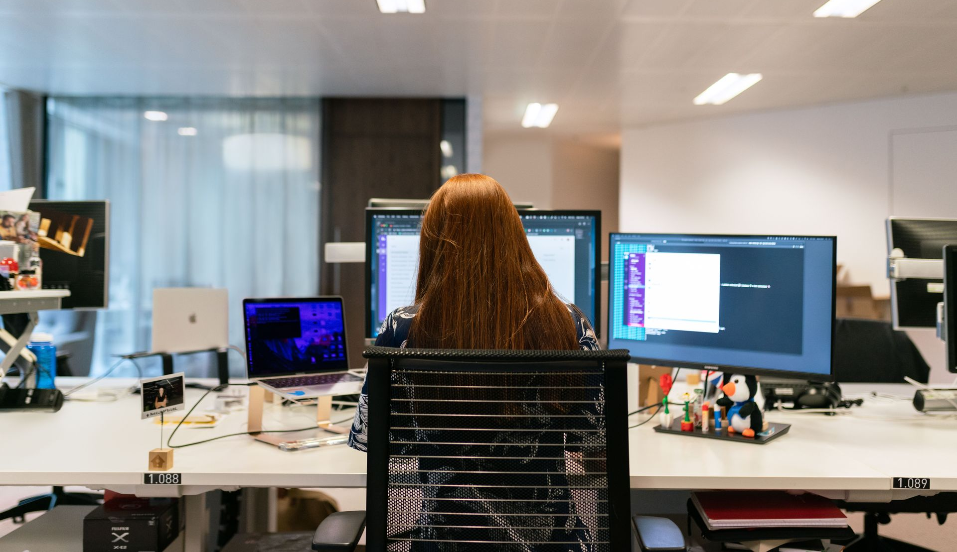 Person seated at a desk in a modern office, working at dual monitors with a laptop nearby