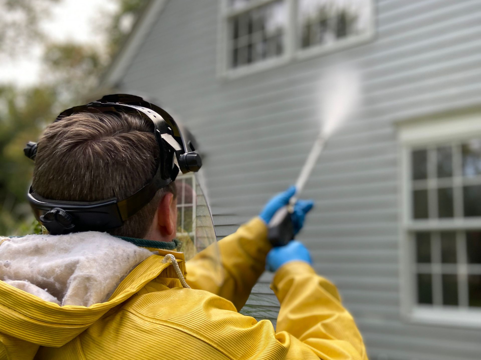 Person in yellow raincoat pressure-washing a house exterior with a blue wand