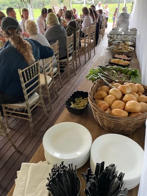 A Group Of People Are Sitting At A Long Table With Plates And Baskets Of Food — Tru Blu Catering In Port Macquarie, NSW