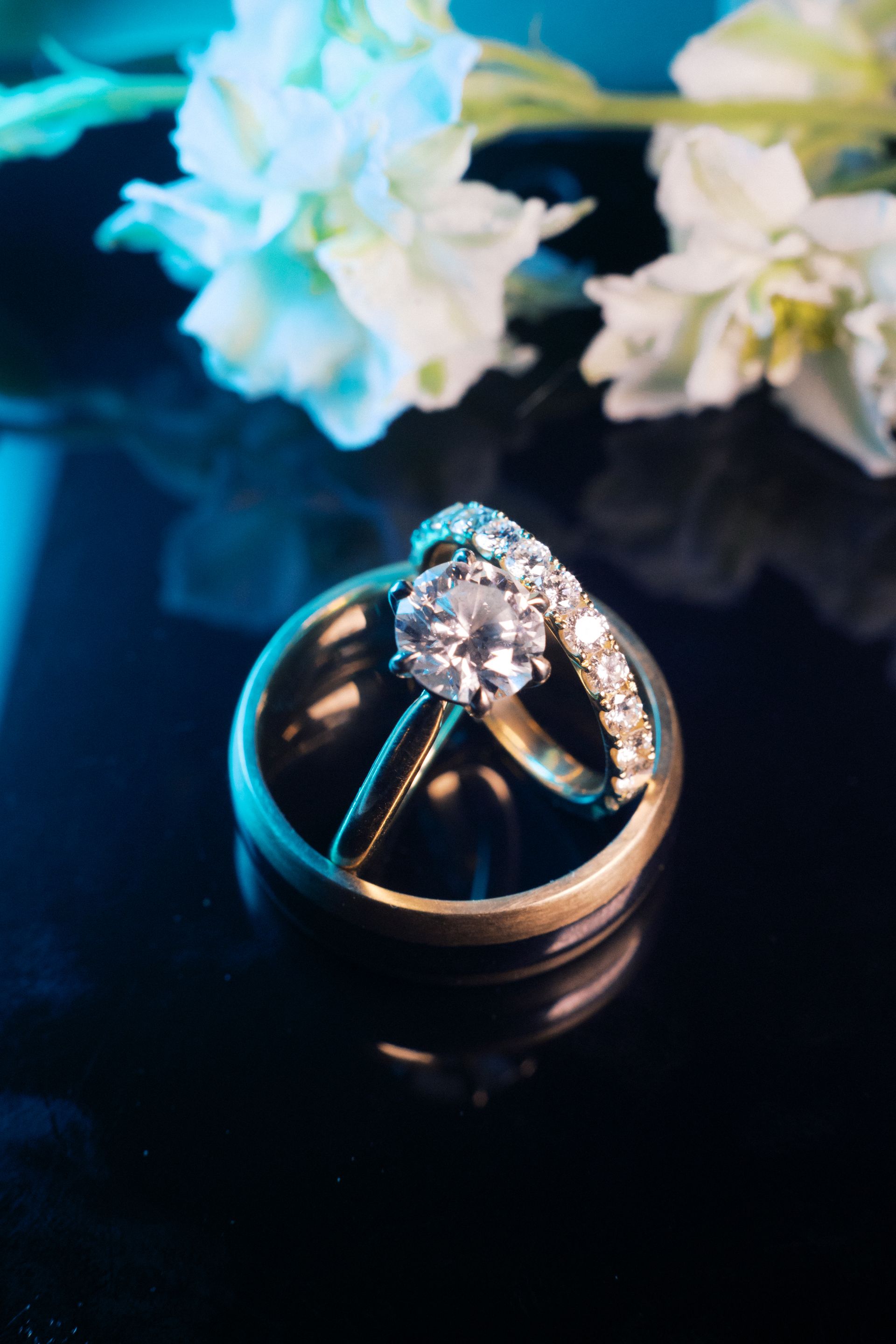 Two wedding rings on a dark surface with white flowers blurred in the background