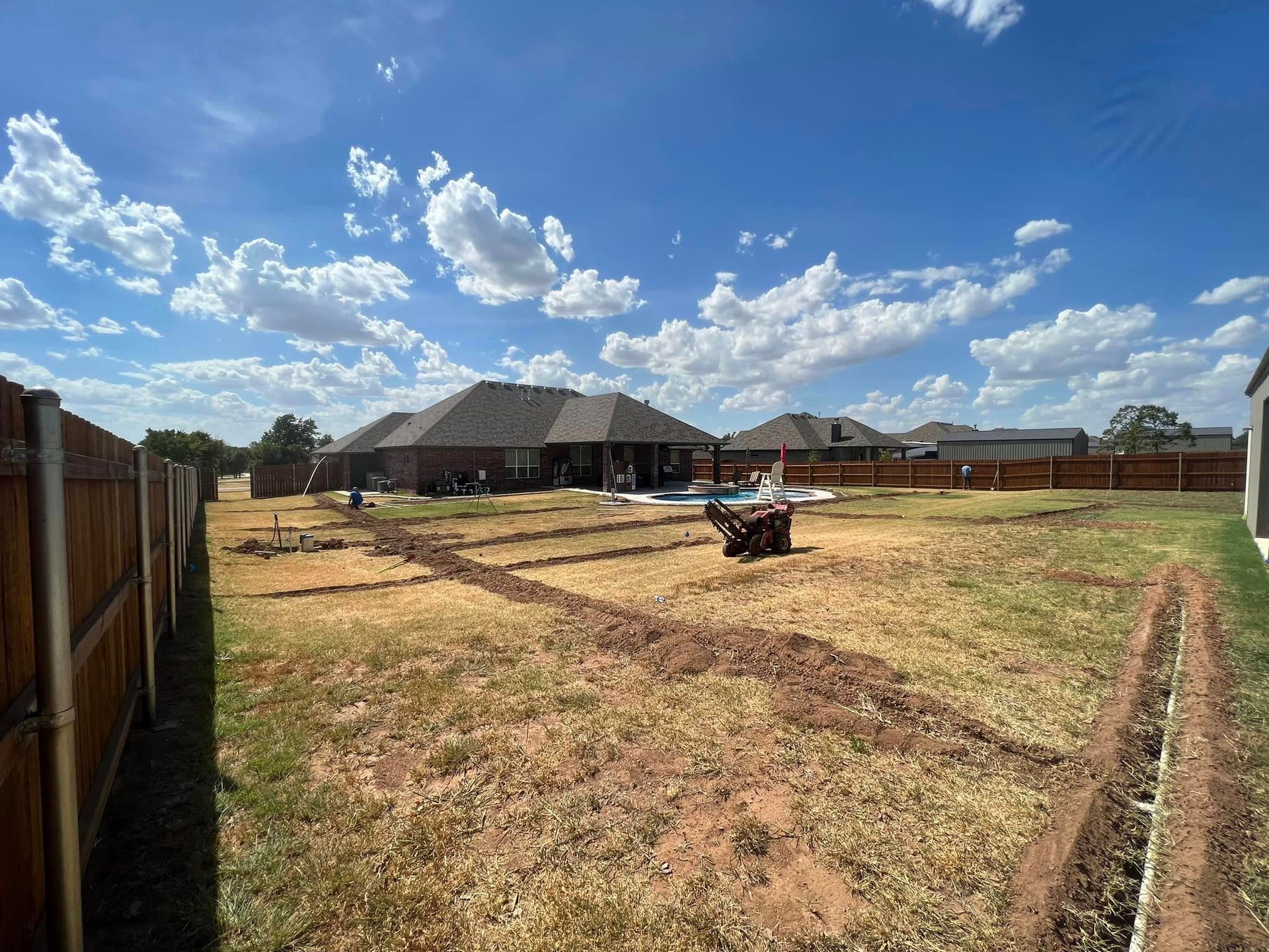 Backyard with trenches being dug for landscaping, under a blue sky with clouds.