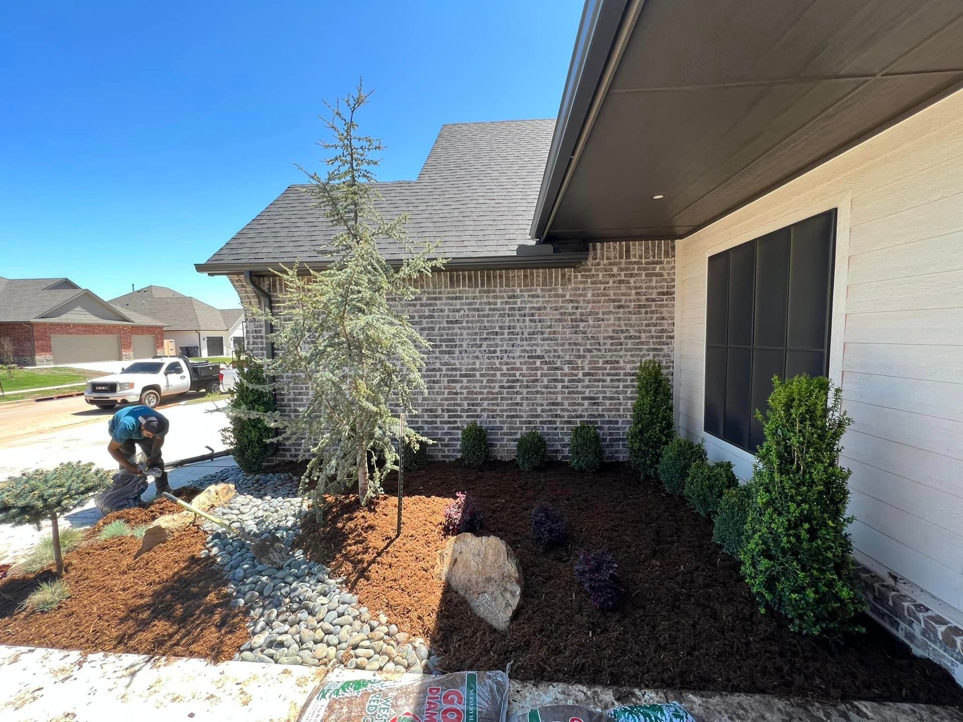 Landscaper working on front yard with new mulch, rocks, and plants. Brick home with blue sky in background.