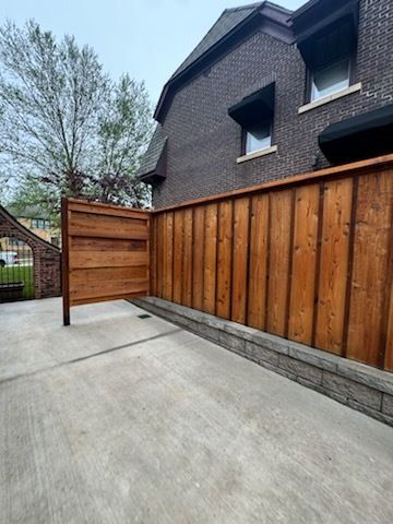 Wooden fence bordering a concrete patio, next to a brick house with awnings.